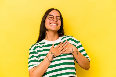 Sarı arka planda izole edilmiş genç İspanyol kadın gülüyor, ellerini kalbe koyuyor, mutluluk kavramını koruyor..