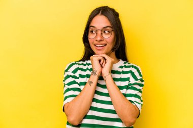 Genç İspanyol kadın sarı arka planda izole edilmiş şans için dua ediyor, şaşırmış ve ağzı açık bir şekilde önüne bakıyor..