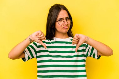 Sarı arka planda izole edilmiş genç İspanyol kadın baş parmağını aşağı gösteriyor, hayal kırıklığı konsepti..