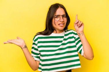 Sarı arka planda izole edilmiş genç İspanyol kadın elinde bir ürün tutuyor..