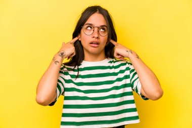 Sarı arka planda izole edilmiş, parmaklarıyla kulaklarını kapatan genç İspanyol kadın, gürültülü bir ortam yüzünden stresli ve çaresiz..