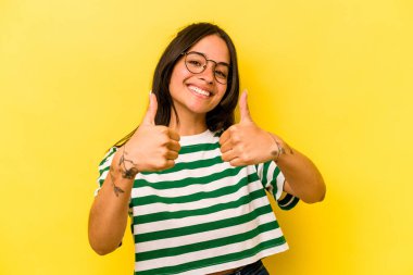 Sarı arka planda izole edilmiş genç İspanyol kadın iki parmağını da kaldırıyor, gülümsüyor ve kendinden emin..