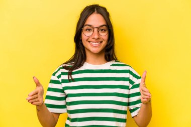 Sarı arka planda izole edilmiş genç İspanyol kadın iki eliyle bir şey tutuyor, ürün sunumu..