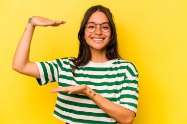 Sarı arka planda izole edilmiş genç İspanyol kadın işaret parmaklarıyla küçük bir şey tutuyor, gülümsüyor ve kendinden emin..
