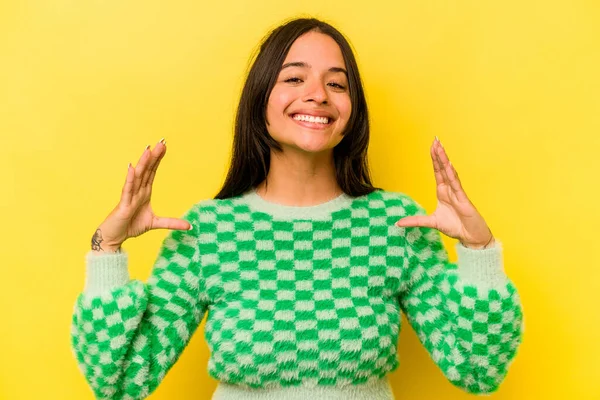 Sarı arka planda izole edilmiş genç İspanyol kadın elinde avuç içi bir şeyle kameraya sunuyor..