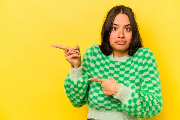 Sarı arka planda izole edilmiş genç İspanyol kadın işaret parmağıyla fotokopi alanını işaret ederek şok oldu..