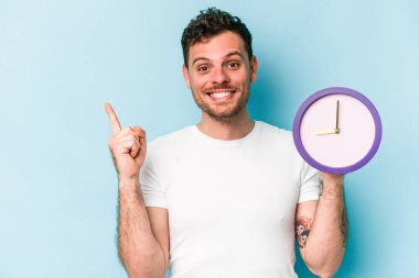 Elinde mavi arka planda izole edilmiş bir saat tutan genç beyaz bir adam gülümsüyor ve boş alanda bir şeyler gösteriyor..