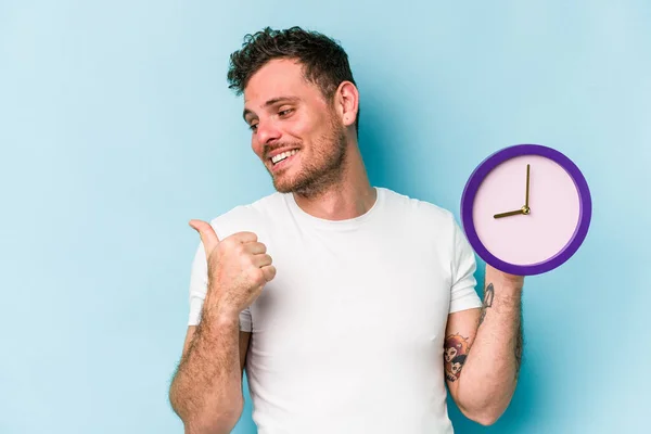 Young Caucasian Man Holding Clock Isolated Blue Background Points Thumb — Fotografia de Stock