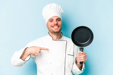 Kafkasyalı genç aşçı elinde uçan tavayla mavi arka planda izole edilmiş gömlek fotokopi alanını işaret eden gururlu ve kendinden emin.