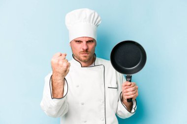 Kafkasyalı genç aşçı elinde uçan tavayla mavi arka planda izole edilmiş. Kameraya yumruk, agresif yüz ifadesi gösteriyor..