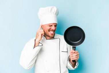 Kafkasyalı genç aşçı elinde uçan tavayla mavi arka planda izole edilmiş kulakları elleriyle kapatıyor..