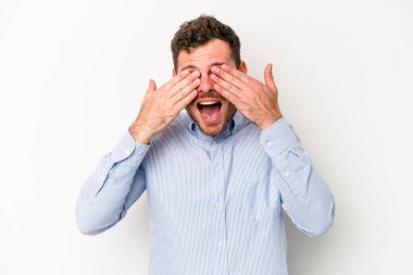 Beyaz arka planda soyutlanmış beyaz tenli genç bir adam elleriyle gözlerini kapatıyor, geniş bir gülümsemeyle bir sürpriz bekliyor..