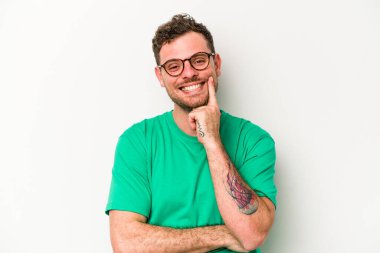 Beyaz arka planda izole olmuş genç beyaz bir adam gülümsüyor ve kendinden emin, çenesine elleriyle dokunuyor..