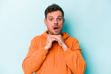 Kafkasyalı genç adam mavi arka planda izole edilmiş şans için dua ediyor, şaşırmış ve ağzını açıp önüne bakıyor..