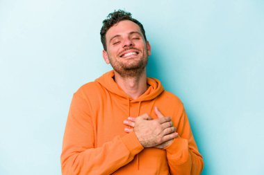 Kafkasyalı genç adam mavi arka planda izole olmuş gülüyor, elini yüreğine koyuyor, mutluluk kavramını koruyor..