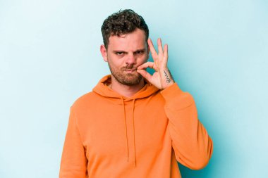 Genç beyaz adam mavi arka planda izole edilmiş ve parmakları dudaklarında bir sır saklıyor..