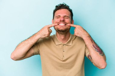 Mavi arka planda izole edilmiş genç beyaz adam gülümsüyor, parmaklarını ağıza doğrultuyor..