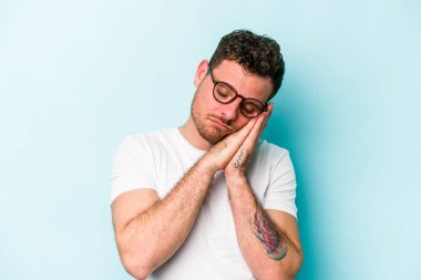 Mavi arka planda izole edilmiş genç beyaz adam esniyor. Ağzı eliyle kapatan yorgun bir hareket sergiliyor..