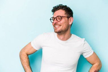 Mavi arka planda izole edilmiş genç beyaz adam mutlu bir şekilde gülüyor ve ellerini karnının üstünde tutarak eğleniyor..