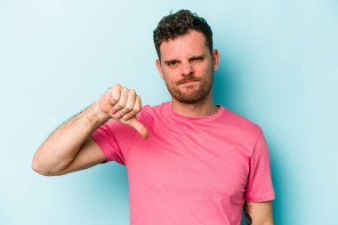 Mavi arka planda izole genç beyaz adam baş parmağını aşağı gösteriyor, hayal kırıklığı konsepti.