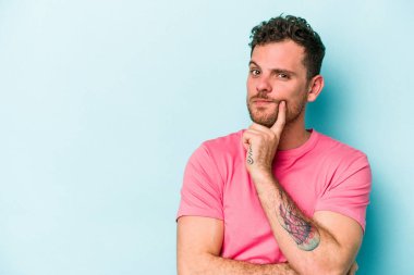 Mavi arka planda izole edilmiş genç beyaz adam şüpheli ve şüpheci bir ifadeyle yana bakıyor..