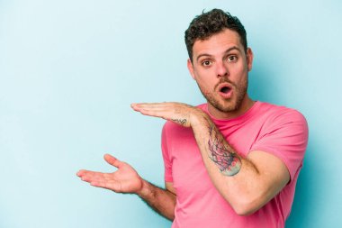 Mavi arka planda izole genç beyaz adam şok ve hayret içinde elleri arasında bir kopya boşluk tutuyor.