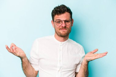 Kafkasyalı genç adam mavi arka planda izole edilmiş sorgulama jestinde şüphe ediyor ve omuz silkiyor..