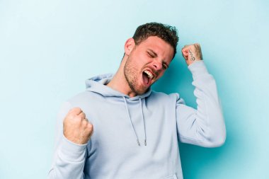 Kafkasyalı genç adam mavi arka planda izole edilmiş kaygısız ve heyecanlı tezahüratlar yapıyor. Zafer konsepti.
