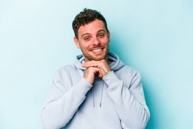 Mavi arka planda izole edilmiş genç beyaz adam ellerini çenesinin altında tutuyor, mutlu bir şekilde yana bakıyor..