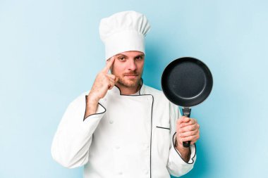 Kafkasyalı genç aşçı elinde uçan bir tavayla mavi arka planda izole edilmiş, parmağıyla tapınağı işaret ediyor, düşünüyor ve bir göreve odaklanıyor..