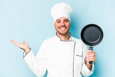 Kafkasyalı genç aşçı elinde uçan tavayla mavi arka planda izole edilmiş bir şekilde avuçta fotokopi alanı ve belinde bir el daha gösteriyor..