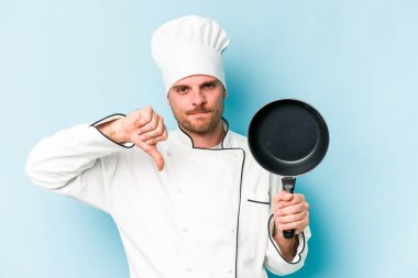 Kafkasyalı genç aşçı elinde uçan tavayla mavi arka planda izole edilmiş. Sevimsiz bir hareket gösteriyor. Anlaşmazlık konsepti.