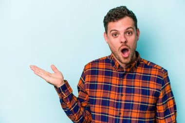 Mavi arka planda izole edilmiş genç beyaz adam, avuç içi fotokopi alanını elinde tutmaktan etkilenmiş..