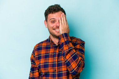 Mavi arka planda izole edilmiş genç beyaz adam yüzünün yarısını palmiye ile kaplayarak eğleniyor..