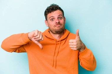 Kafkasyalı genç adam mavi arka planda izole edilmiş, baş parmağını kaldırıyor, zor seçim konsepti.