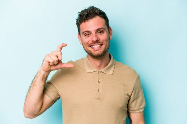 Kafkasyalı genç adam mavi arka planda izole edilmiş. Elinde işaret parmaklarıyla küçük bir şey tutuyor, gülümsüyor ve kendinden emin..