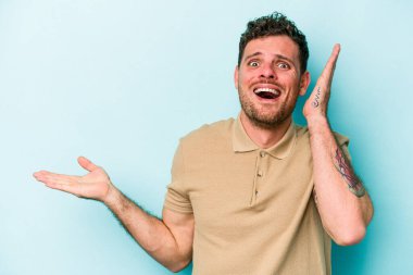 Mavi arka planda izole edilmiş genç beyaz bir adam avucunun içinde fotokopi alanı tutuyor, el üstünde tutuyor. Şaşırmış ve mutlu..