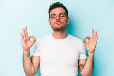 Mavi arka planda izole edilmiş genç beyaz adam yorucu bir günün ardından rahatlıyor. Yoga yapıyor..