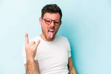 Mavi arka planda izole edilmiş genç beyaz adam parmaklarıyla kaya hareketi gösteriyor.