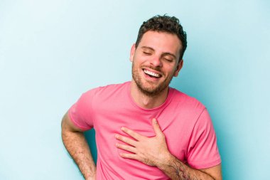 Kafkasyalı genç adam mavi arka planda izole olmuş gülüyor, elini yüreğine koyuyor, mutluluk kavramını koruyor..