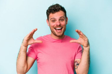 Kafkasyalı genç adam mavi arka planda izole edilmiş. Elinde avuç içi olan bir şey var ve kameraya sunuyor..