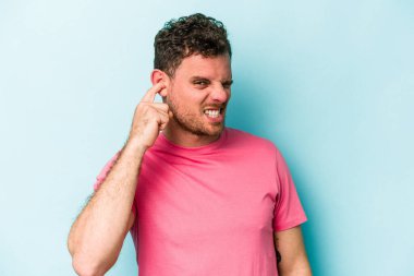 Genç beyaz adam mavi arka planda izole edilmiş parmaklarıyla kulaklarını kapatıyor, gürültülü bir ortam tarafından stresli ve umutsuz..
