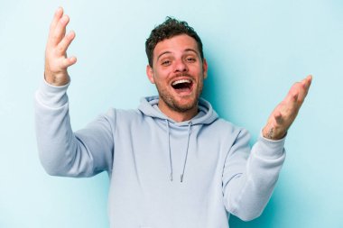 Mavi arka planda izole edilmiş genç beyaz bir adam zaferi ya da başarıyı kutluyor, şaşırıyor ve şok oluyor..