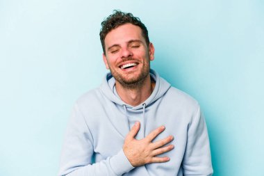 Mavi arka planda izole olmuş genç beyaz adam yüksek sesle gülüyor ve elini göğsüne koyuyor..