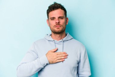 Kafkasyalı genç adam mavi arka planda izole edilmiş yemin ediyor, elini göğsüne koyuyor..
