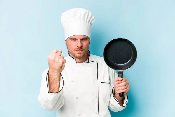年轻的高加索厨师手持飞盘 与蓝色背景隔离 对着相机显示拳头 咄咄逼人的面部表情 — 图库照片