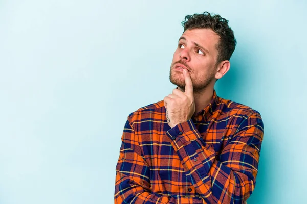 Mavi arka planda izole edilmiş genç beyaz adam şüpheli ve şüpheci bir ifadeyle yana bakıyor..
