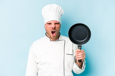 Kafkasyalı genç aşçı elinde uçan tavayla mavi arka planda izole edilmiş kızgın ve agresif çığlıklar atıyor..