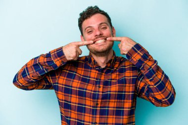 Mavi arka planda izole edilmiş genç beyaz adam gülümsüyor, parmaklarını ağıza doğrultuyor..