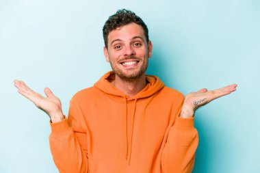 Mavi arka planda izole genç beyaz adam kollarıyla ölçek yapar, mutlu ve kendinden emin hisseder..